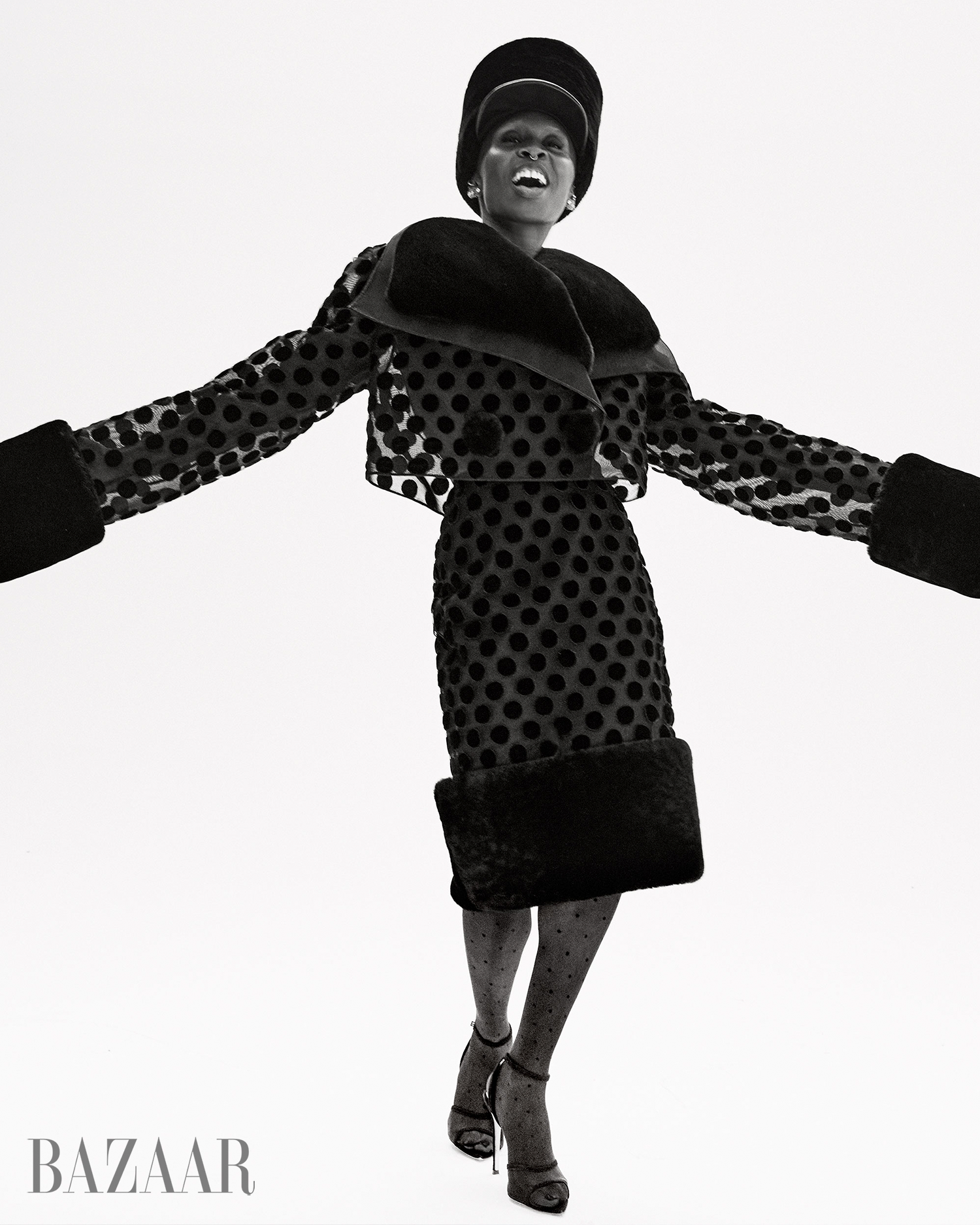 A Black female fashion model in a black polka dot skirt suit, hat, and heels poses in a photo studio with a white backdrop.