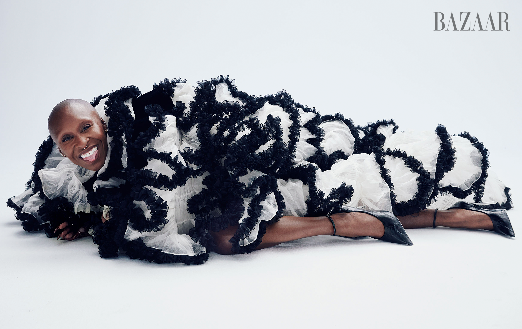 Woman in an elaborate black and white ruffled dress lies on a white surface, sticking her tongue out and wearing black high heels.
