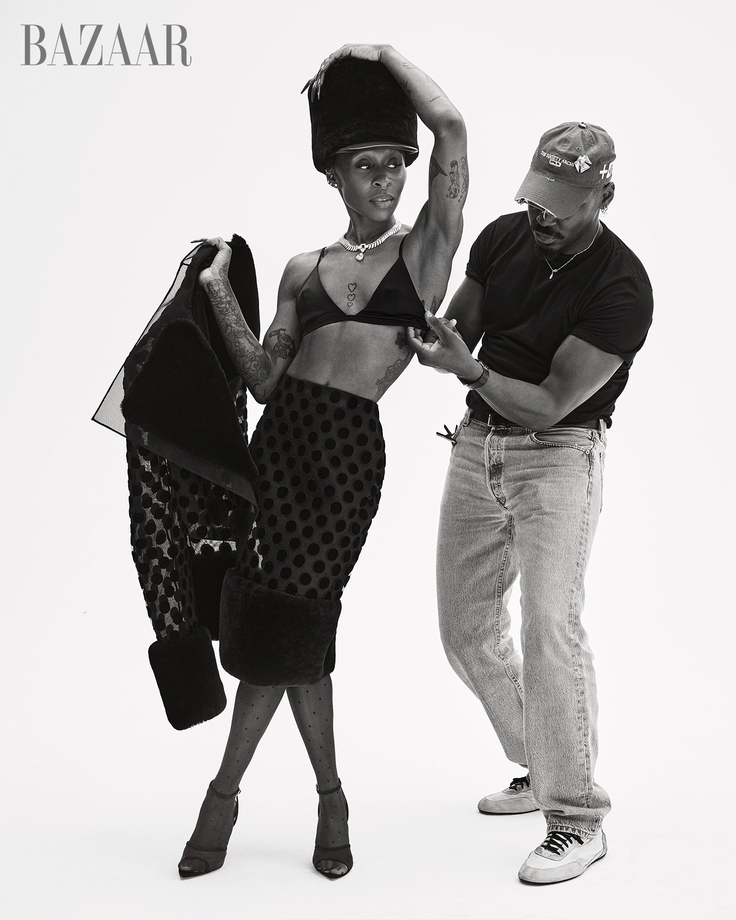Fashion editorial shot of an adult female in a skirt, bralette, and hat posing with a man adjusting her clothing in a studio.
