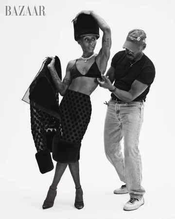 Fashion editorial shot of an adult female in a skirt, bralette, and hat posing with a man adjusting her clothing in a studio.
