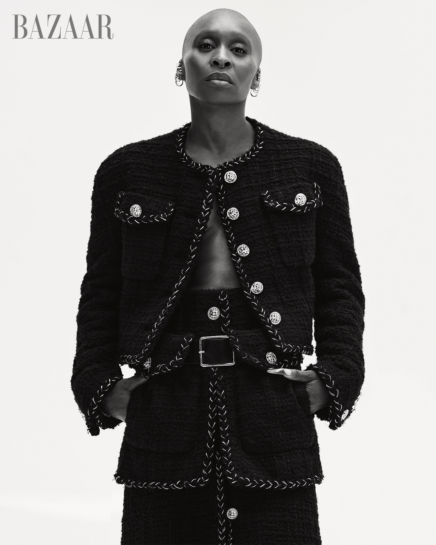Black and white portrait of a bald, dark-skinned woman in a tweed jacket and skirt with a belt, posing against a bright background.