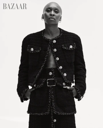 Black and white portrait of a bald, dark-skinned woman in a tweed jacket and skirt with a belt, posing against a bright background.