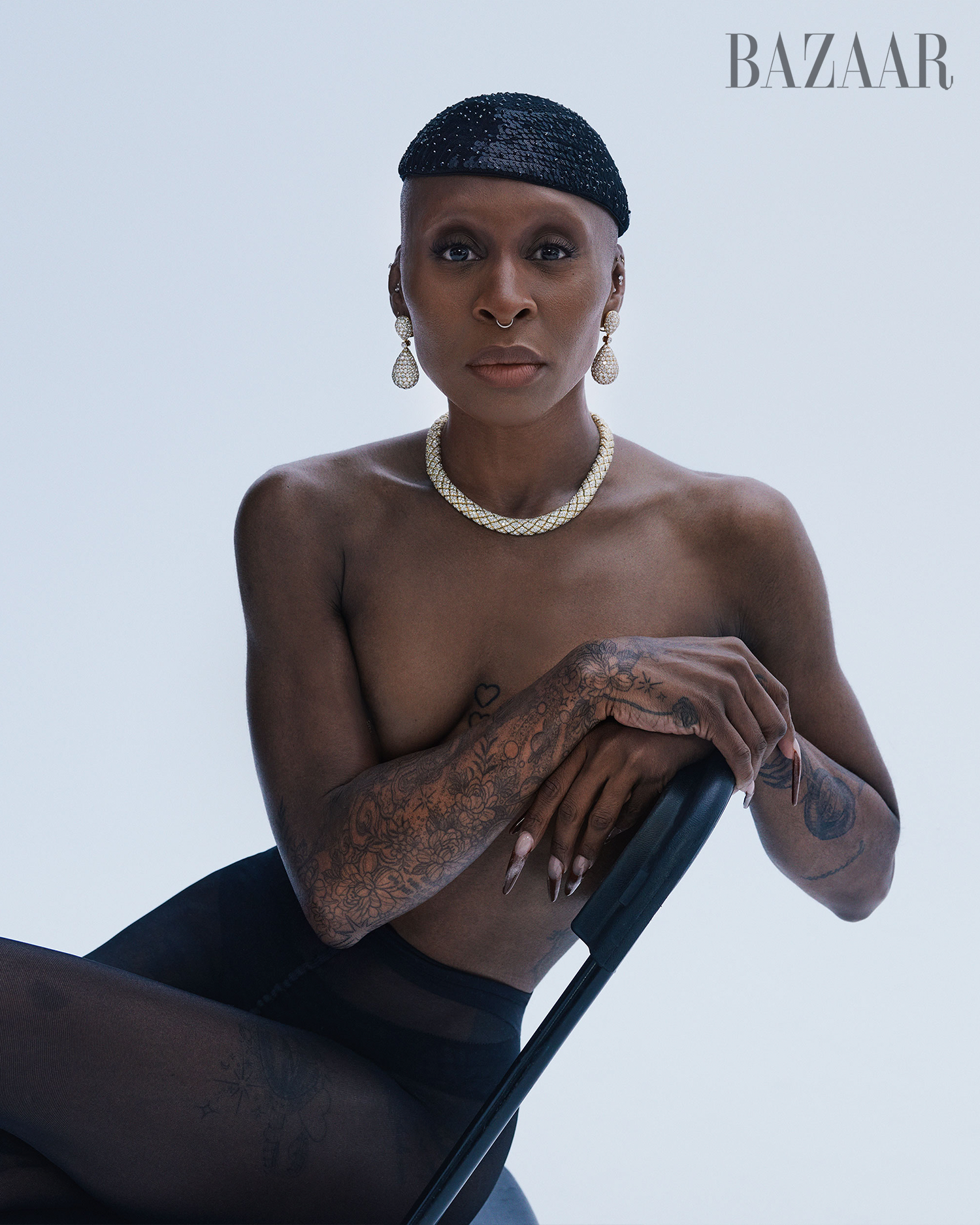 Elegant woman in a black hat, nude shoulders, and pearl jewelry, posing in a bright studio, seated on a black chair for a fashion shot.