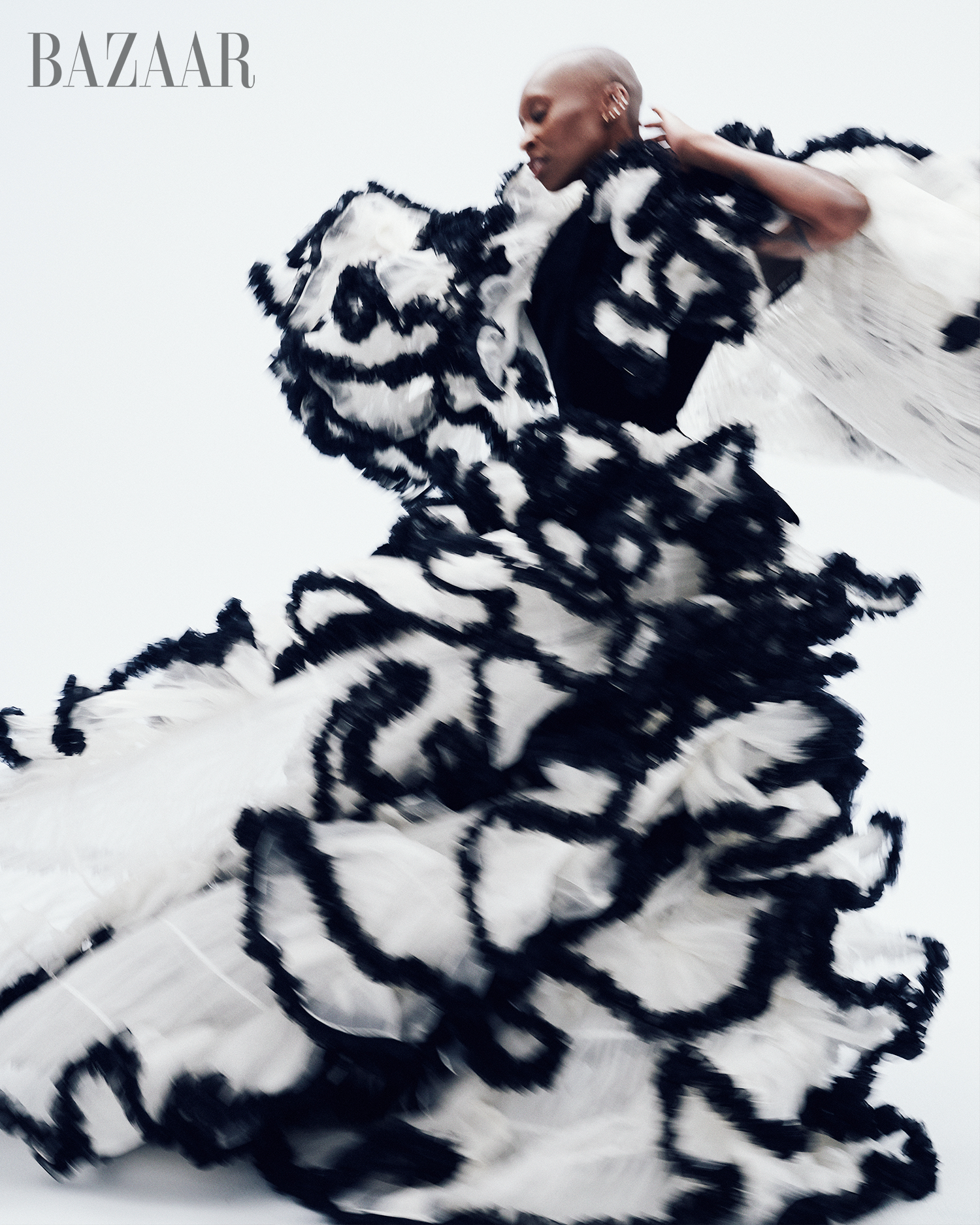 Elegant woman with shaved head wearing black top and black and white ruffled dress on white backdrop, fashion editorial for Bazaar.