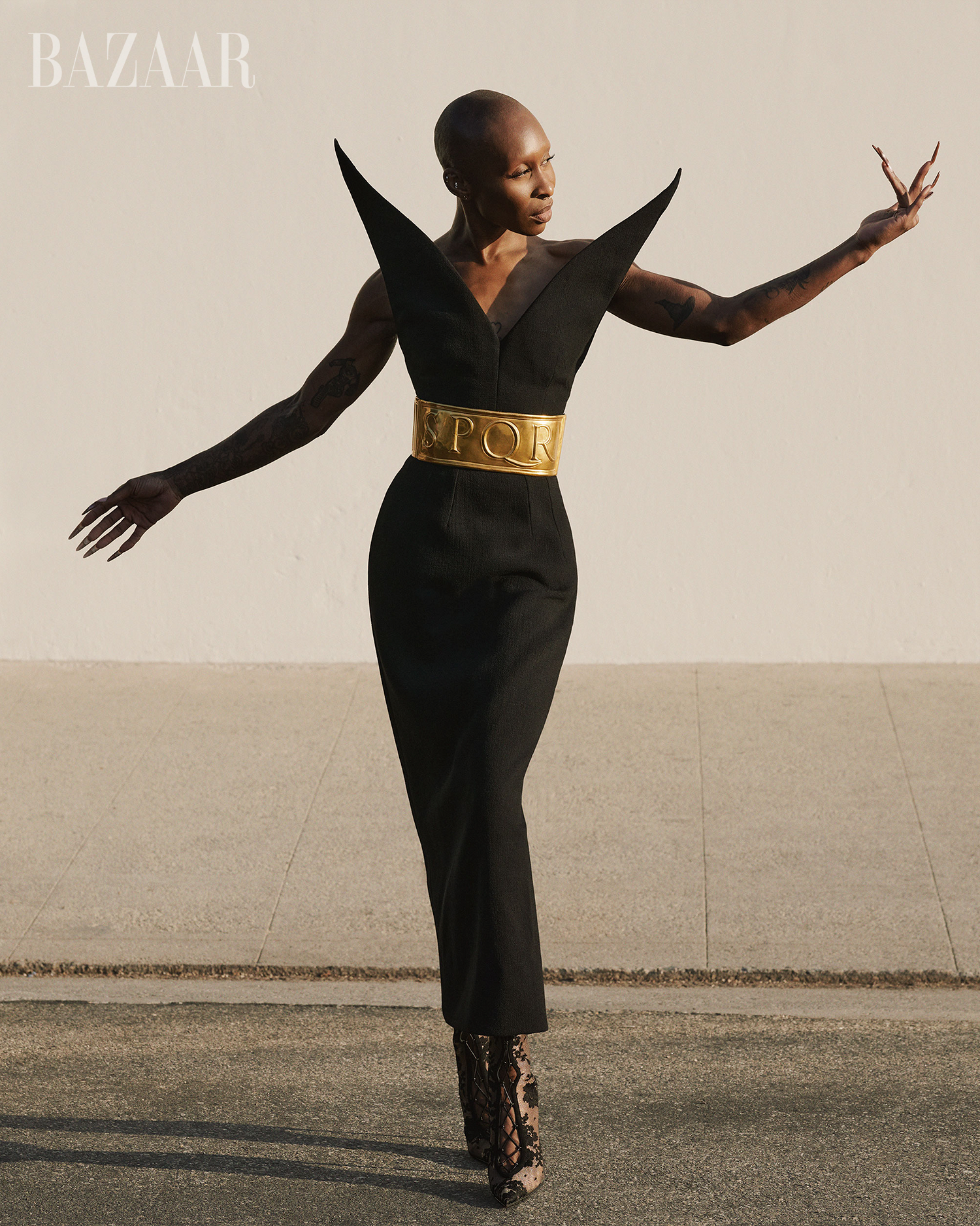 Fashion shot of a Black woman in a black dress with statement shoulders and a gold belt, posing outdoors with arms extended in a street.