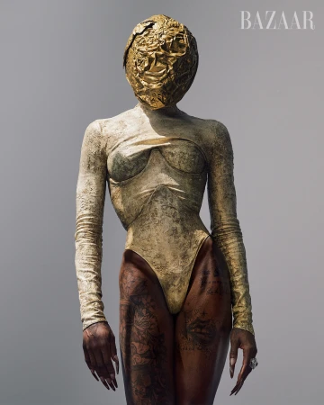 Fashion portrait of a woman in a gold bodysuit and head covering against a neutral background, with the Bazaar logo visible.
