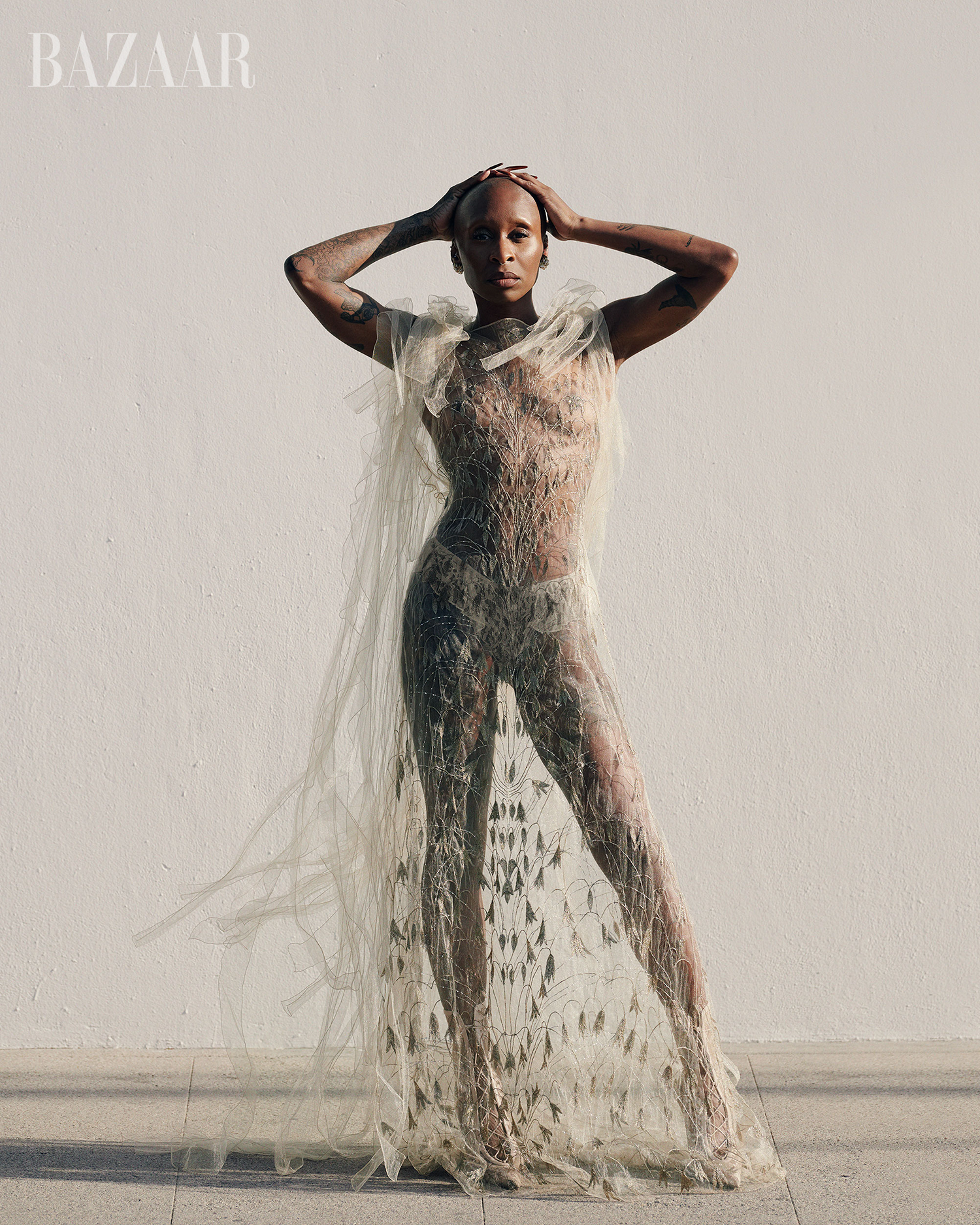 Bald woman posing in sheer couture dress against a white wall in a fashion editorial-style photograph; sun casts shadows on the wall.