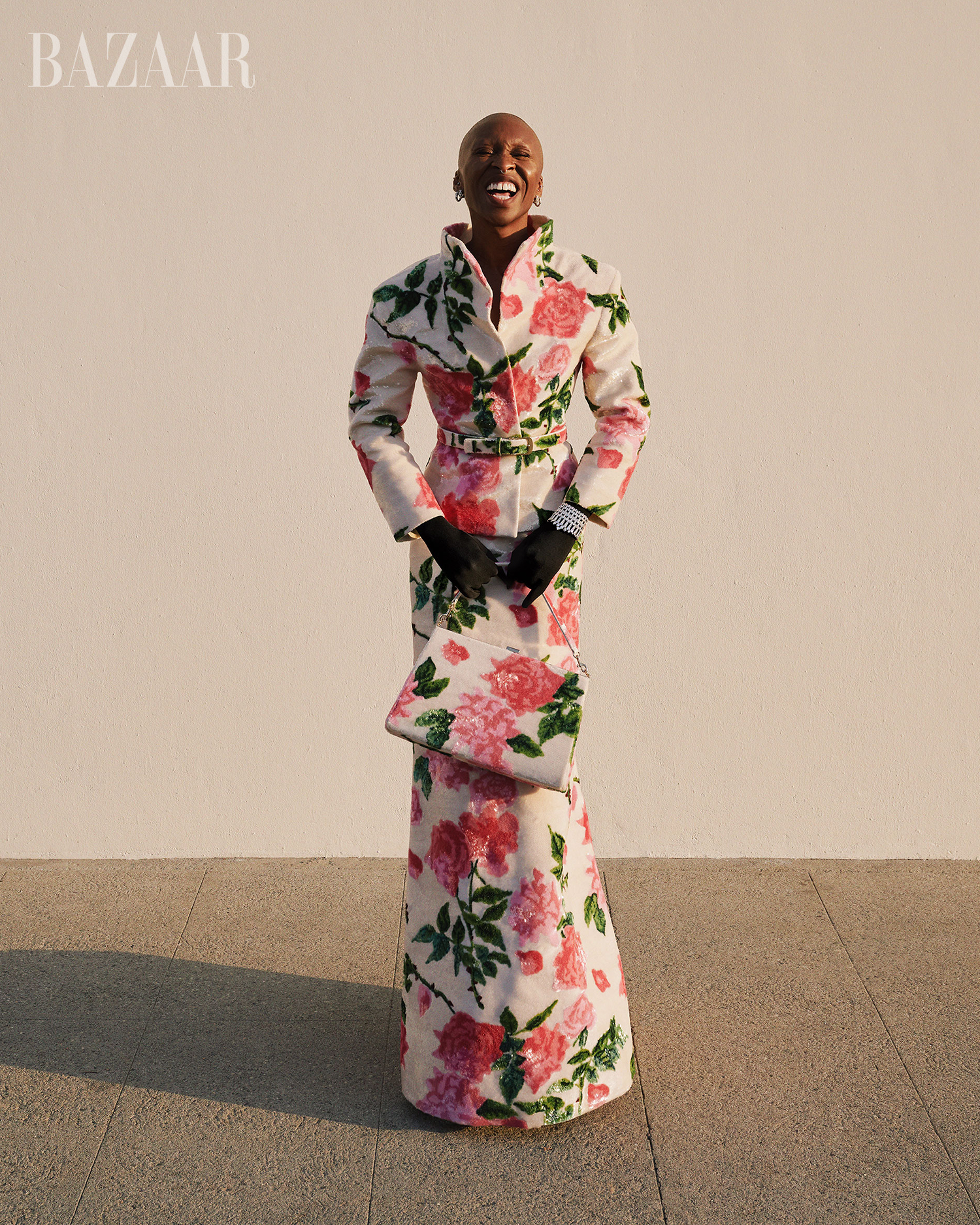 Full-length portrait of an African-descent adult female smiling outdoors, wearing a floral-patterned suit with matching clutch and black gloves.