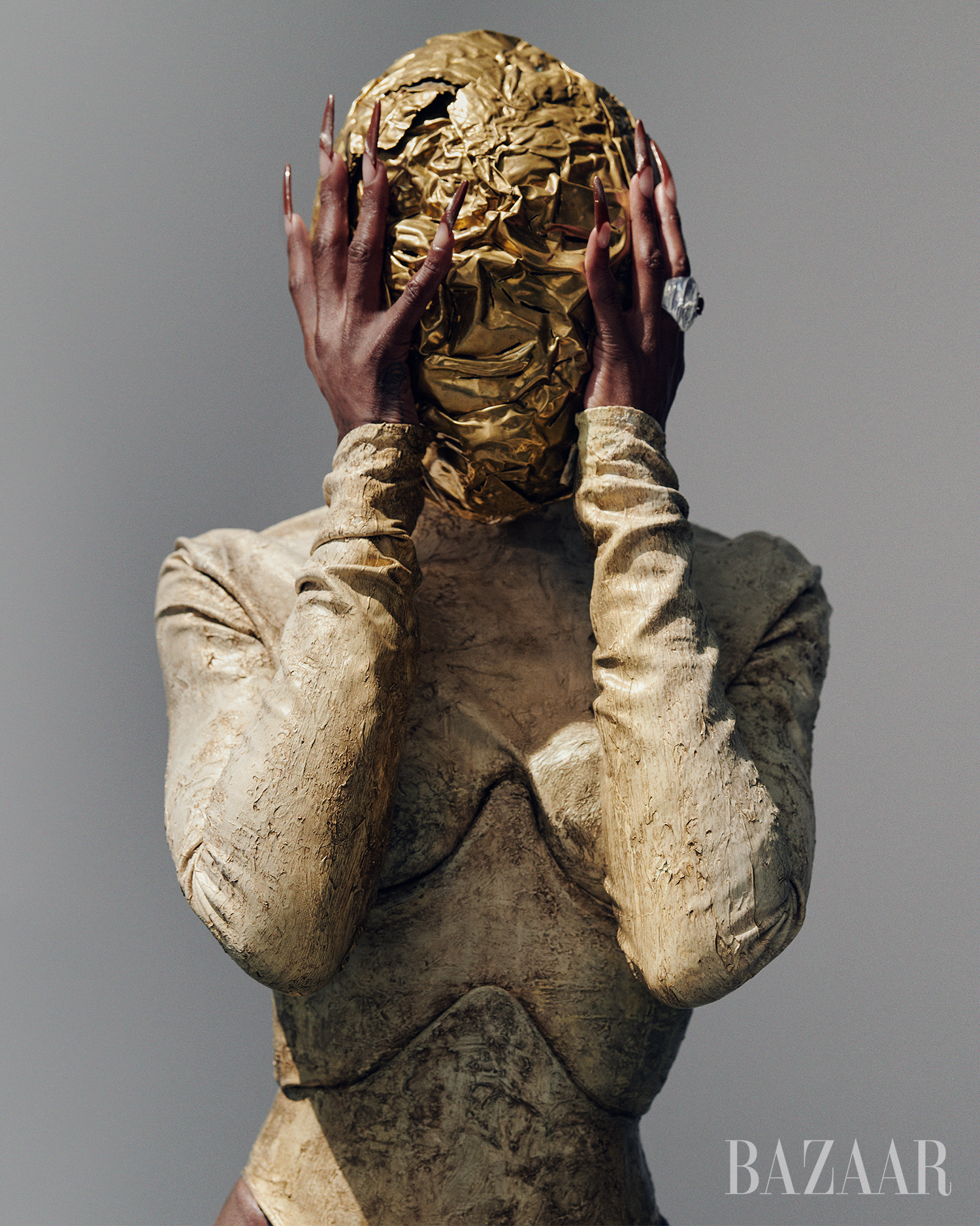 Woman wearing gold textured full bodysuit and head covering, posing with hands near face.  Long fingernails and large ring are visible.