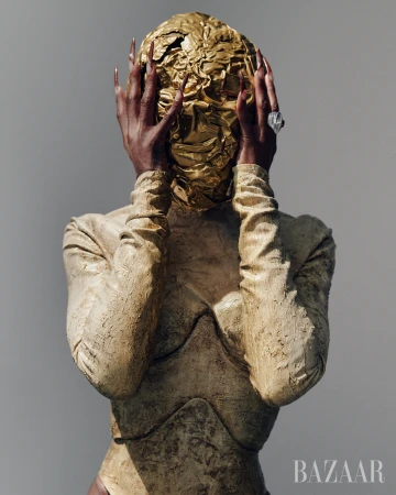 Woman wearing gold textured full bodysuit and head covering, posing with hands near face.  Long fingernails and large ring are visible.