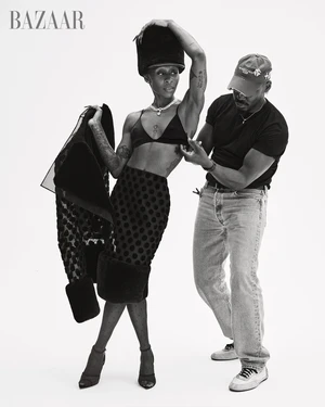 Black and white portrait of Cynthia Erivo posing in a bralette and skirt while a stylist adjusts her outfit on a white background