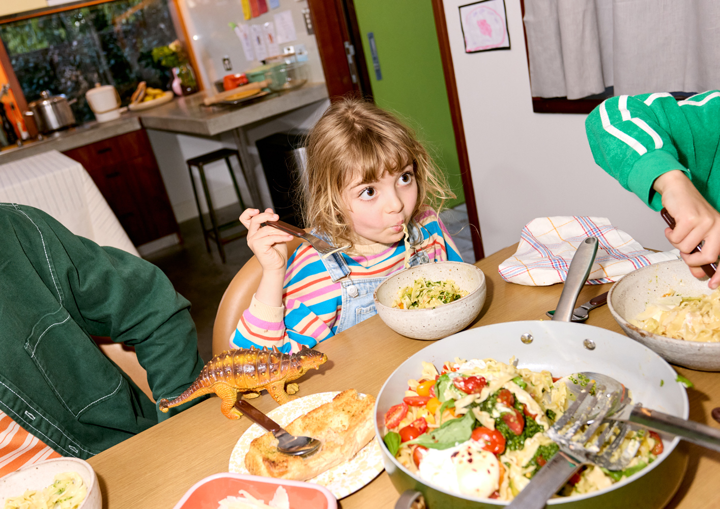 Young girl with blonde hair eating pasta at a table indoors, with other children and a dinosaur toy also visible.