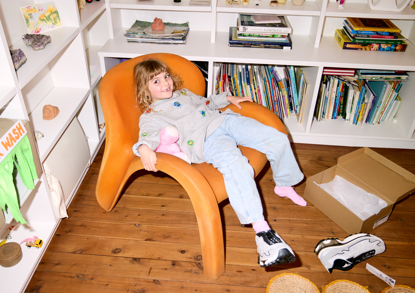 A young child with blond hair is sitting in an orange chair. Shoes are on the floor, and a white bookshelf is visible in the background.