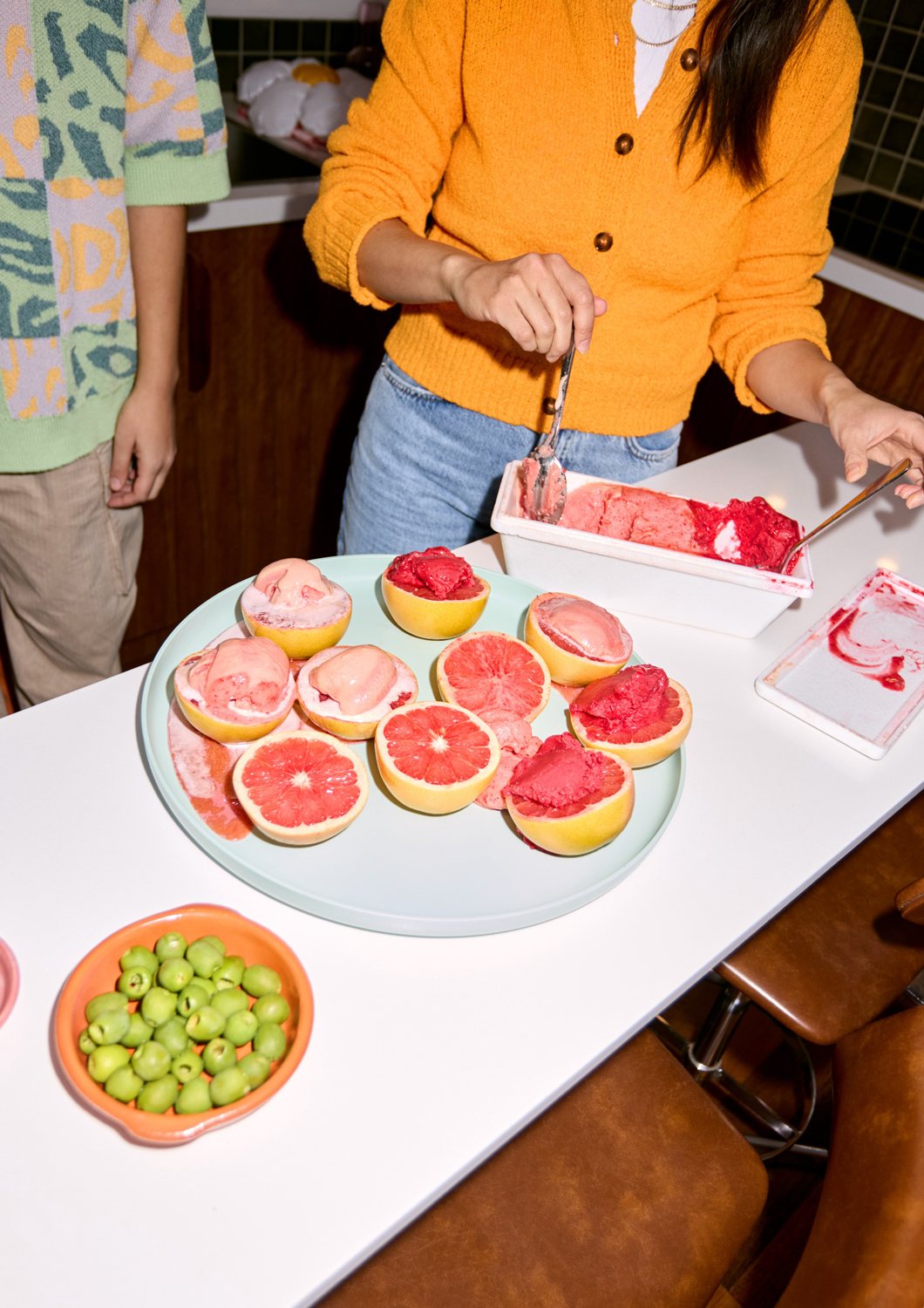 Woman scoops pink sorbet into grapefruit halves on a white table, alongside green candies, with another person and kitchen visible.