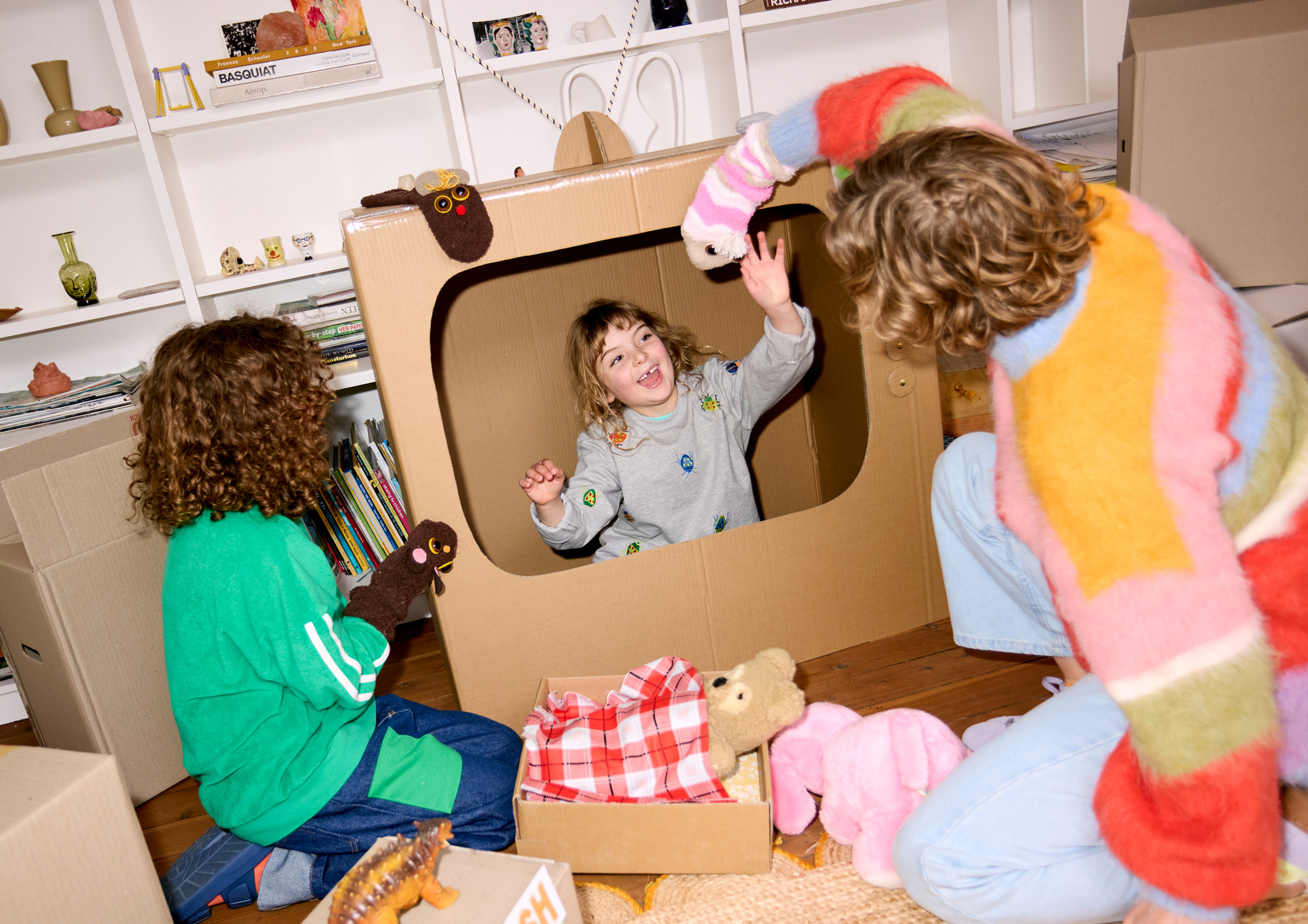 Three children play with a cardboard television and animal puppets indoors in a playfully staged scene featuring props and bright colours.
