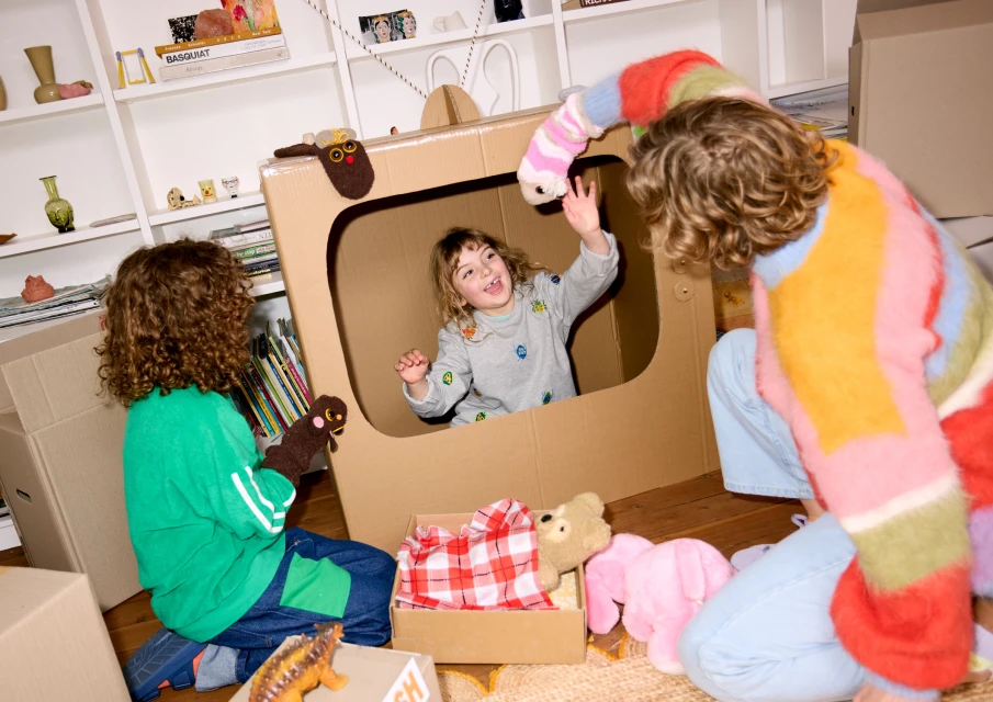 Three children play with a cardboard television and animal puppets indoors in a playfully staged scene featuring props and bright colours.