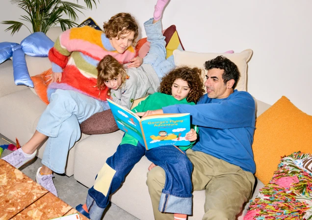 Family of four gathered on a couch, reading a book together in a home setting with cushions and a coffee table. Reading time.