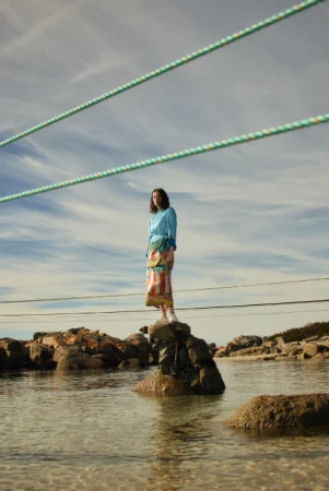 A person stands on a rock in water under an open sky, wearing a blue sweater and patterned skirt. Green lines stretch across the sky.