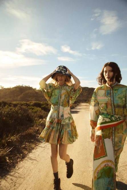 Two women in patterned dresses walk on a dirt road under a bright sky, surrounded by dry vegetation in an outdoor fashion shot.