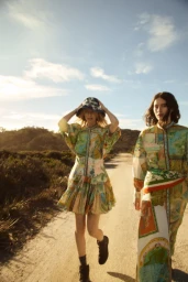 Two female models wearing patterned dresses and hats walking down a dusty road under an open sky
