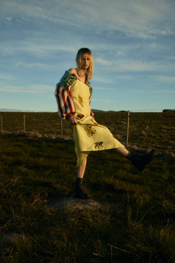 Model standing in a field against a blue sky, wearing a yellow patterned dress with striped sleeves and dark boots.