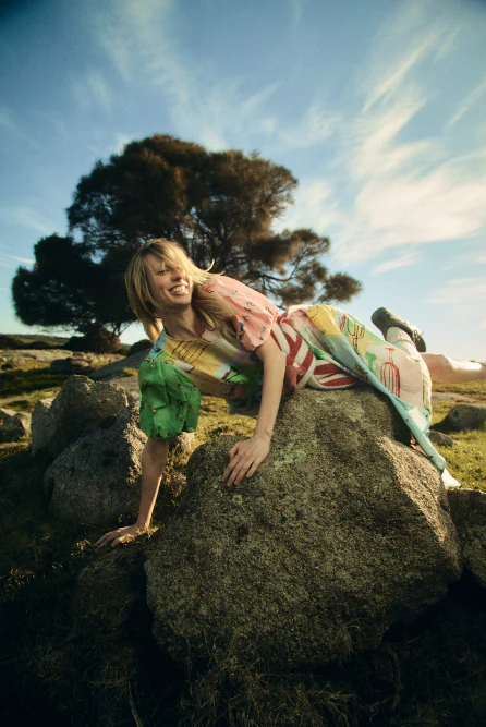 Model wearing a colourful patterned dress climbing over large boulders in an outdoor landscape.