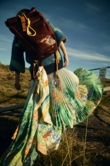 Woman in flowing green dress with floral design carries brown leather backpack on rural grassy hillside under blue sky.