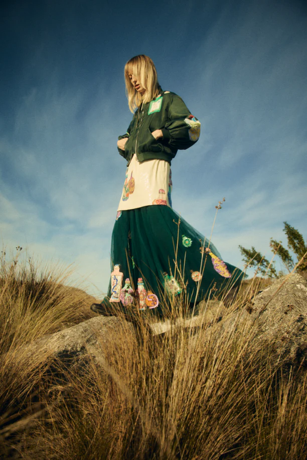 Woman in a green jacket and long printed skirt standing on rocks in a field of tall grass, with a blue sky and clouds backdrop.