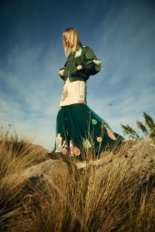 Model standing on a rocky grassy outcrop wearing a dark green bomber jacket and patterned maxi skirt against a bright blue sky.