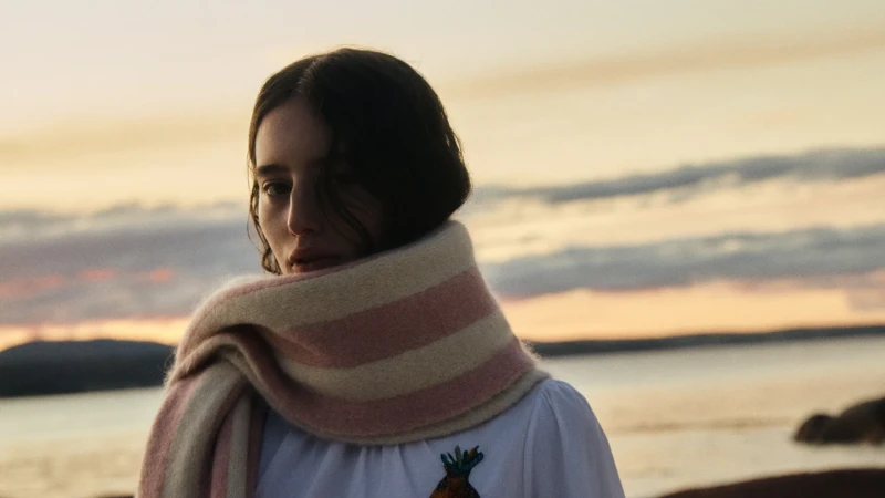 Woman wearing a thick striped scarf wrapped around her neck with a sunset sky and ocean background