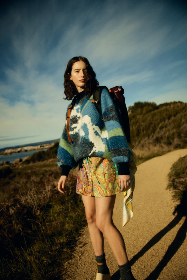 Young woman on a hiking path, wearing a patterned sweater and shorts, with a backpack, on a sunny day with a blue sky.