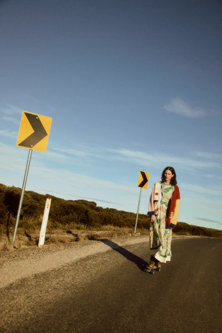 Woman on skateboard on a road with directional road signs under a blue sky. She is wearing a floral dress and cardigan.