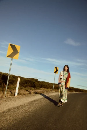 Woman on skateboard on a road with directional road signs under a blue sky. She is wearing a floral dress and cardigan.