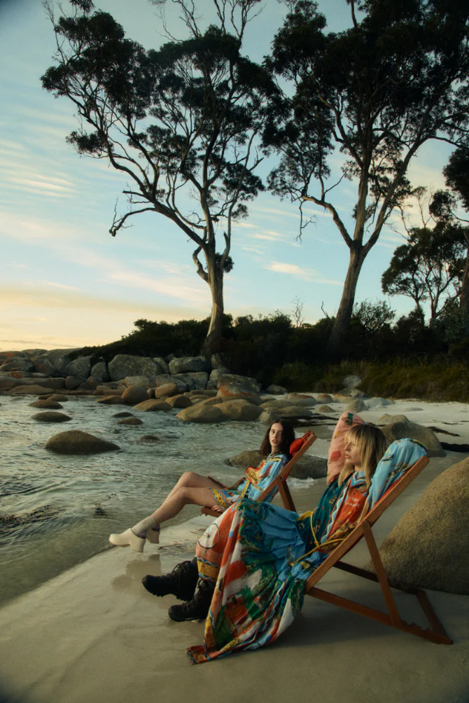 Two women in colourful summer dresses lounge in deck chairs on a sandy beach with trees and rock formations during daytime.
