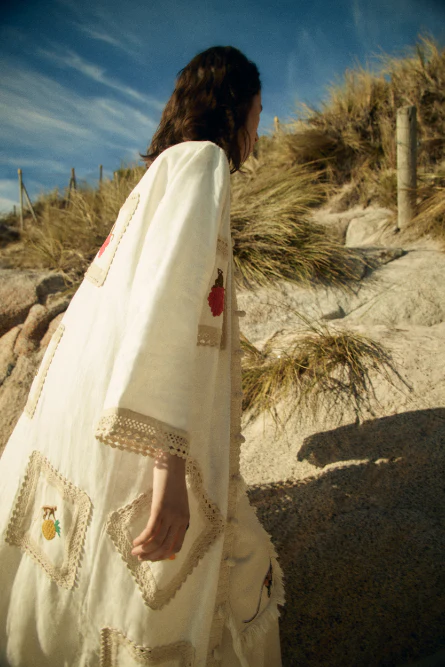 Woman in white embroidered robe walking on a sandy beach with grassy dunes under a bright sky, capturing a serene fashion moment.