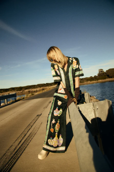 Woman poses in printed shirt and pants on bridge against blue sky and water. Fashion shot highlighting apparel and outdoor location.