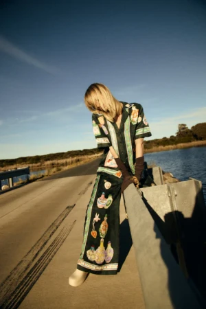 Woman poses in printed shirt and pants on bridge against blue sky and water. Fashion shot highlighting apparel and outdoor location.