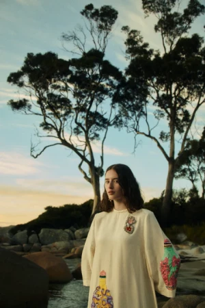 Woman in cream embroidered dress standing on a rocky shore with large trees under an overcast sky for Island Odyssey Fall 24.