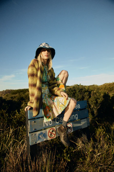A young adult woman with blonde hair wearing a hat and patterned cardigan sits on a signpost that reads 'No Camping' in an outdoor setting.