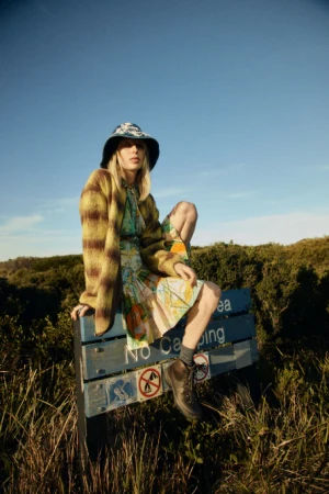 A young adult woman with blonde hair wearing a hat and patterned cardigan sits on a signpost that reads 'No Camping' in an outdoor setting.