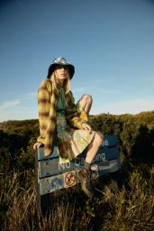 Model seated on a blue sign wearing a patterned dress, fuzzy cardigan, bucket hat, and boots against a clear sky