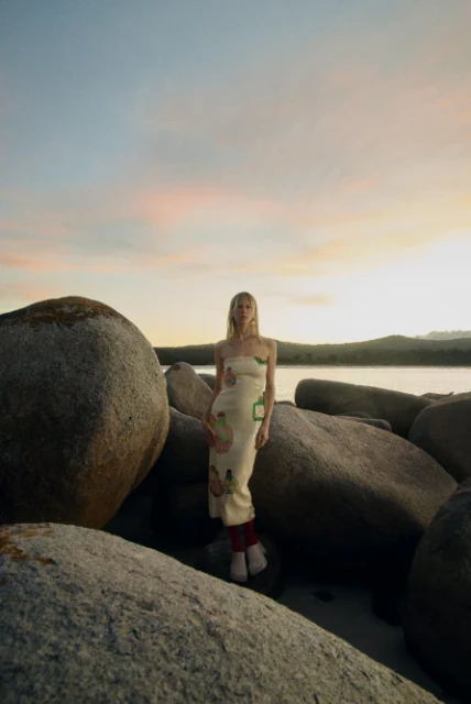Woman poses on rocky beach at sunset wearing a printed dress and boots, framed by large boulders against a pink and blue sky.