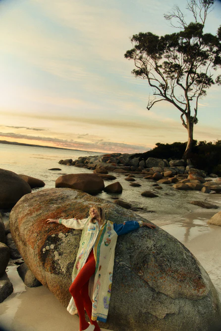 Woman in patterned coat rests on rock at a beach at dusk, featuring calm ocean, sandy shore, and a single tree in the background.