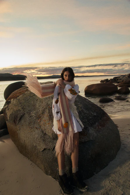 A young woman in a white dress and patterned scarf poses on a beach by a large rock formation at sunset, overlooking the water.