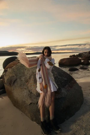 A young woman in a white dress and patterned scarf poses on a beach by a large rock formation at sunset, overlooking the water.