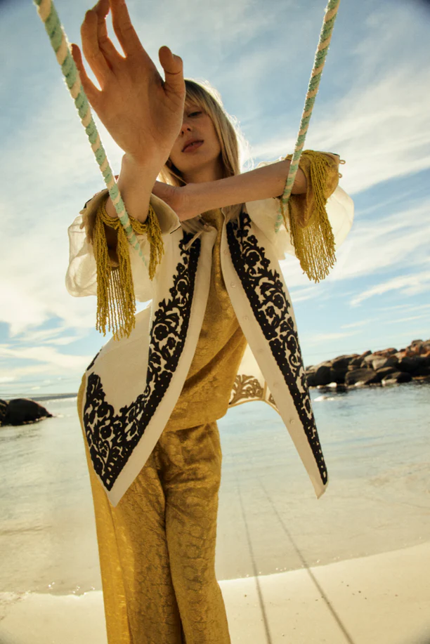 A fair-skinned woman in a decorative vest and patterned pants stands on a beach, holding a rope with a blue sky and ocean in the background.