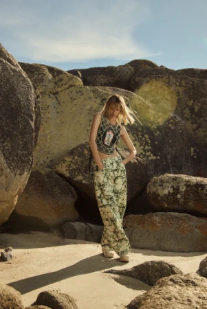 A woman in patterned pants and a crop top poses between large rock formations in an outdoor fashion shot on a sunny day.