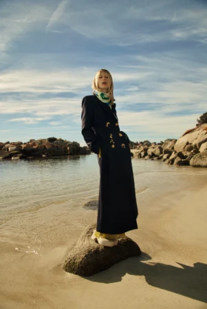A blonde model wearing a dark long coat and patterned scarf standing on a rock on a sandy beach under a blue sky.