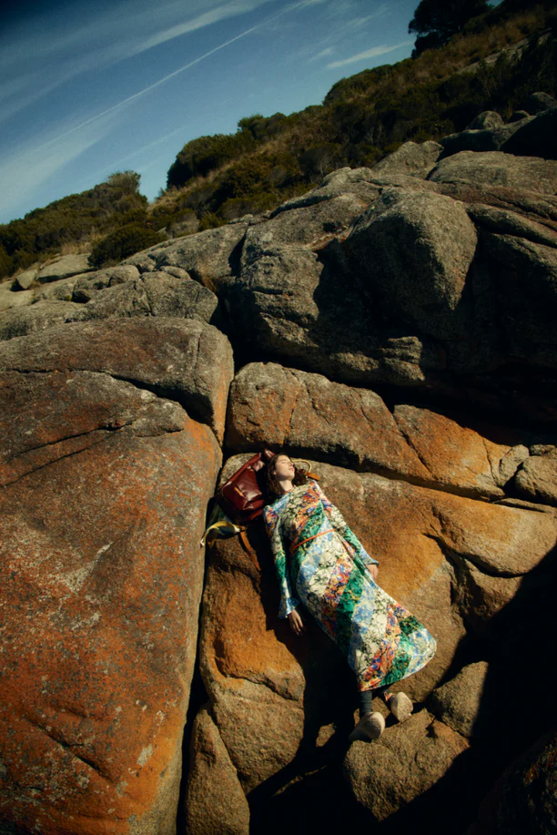 Woman in floral dress lies on coastal rocks with brown leather bag. A tranquil outdoor scene on a sunny day near vegetation.