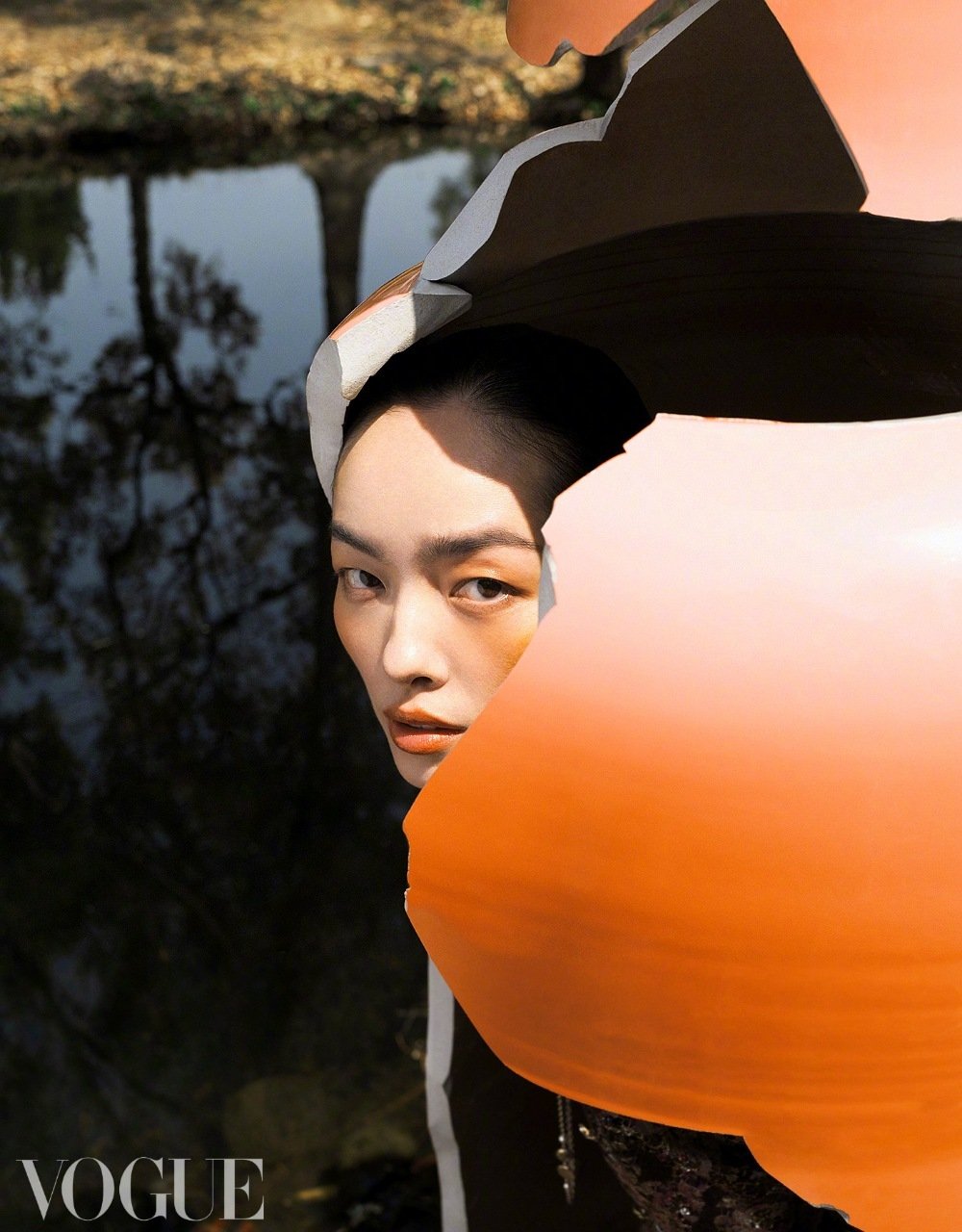 Portrait of an Asian woman with dark hair and light skin partially obscured by an abstract sculpture in an outdoor setting with water.